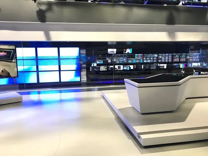 The end of Al Araby TV's news desk beside a video wall, looking through glass into the gallery's monitor walls and operator positions.