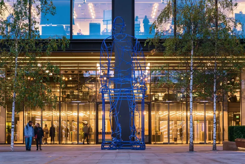 The glass entrance of Amazon's LHR16 office in London at dusk, with a large blue wireframe sculpture of a seated figure out front.