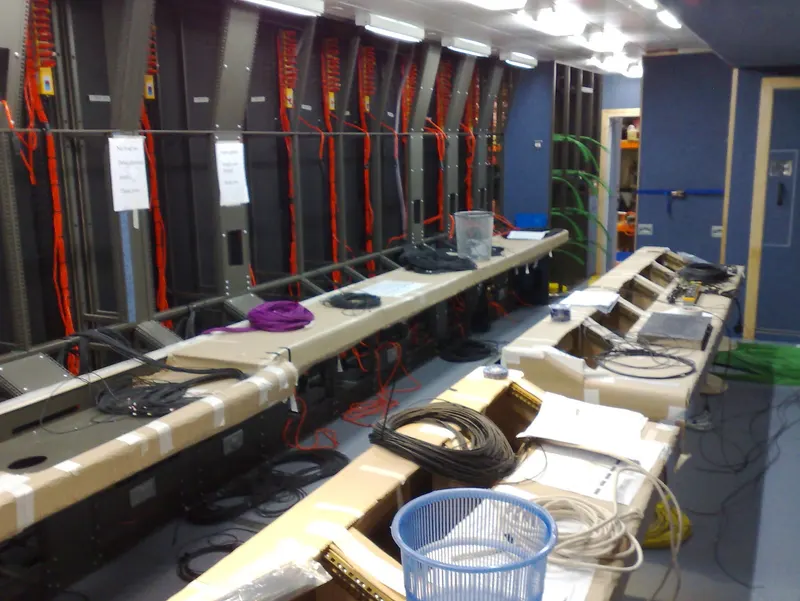 The production area inside an O21 Television OB truck during fit-out, with desks still protected in cardboard and rack frames wired with orange cable behind them.