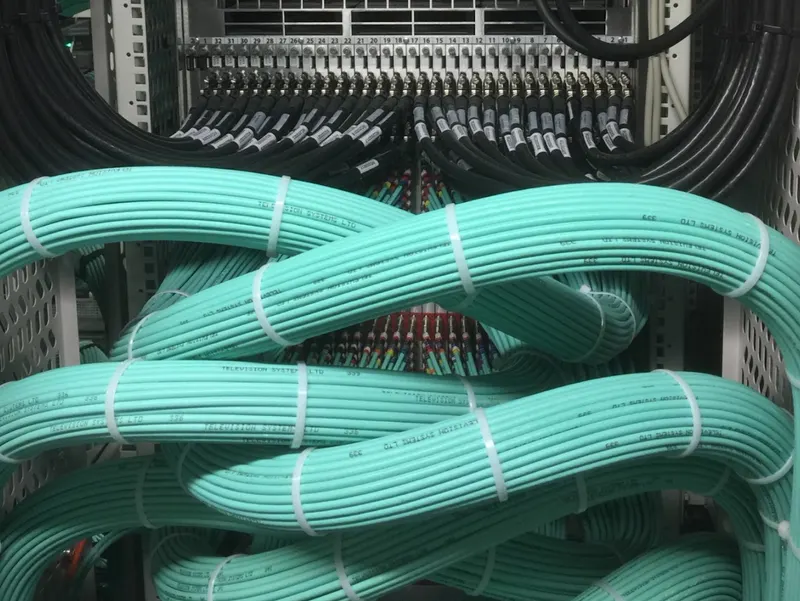 Looped bundles of teal multicore cable dressed beneath a row of labelled coaxial connections in IMG's Stockley Park data centre.