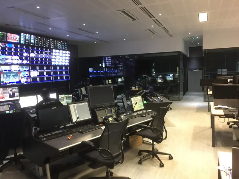 A row of engineering desks in IMG's Stockley Park master control room, with the monitor wall on the left and glass-walled suites behind.