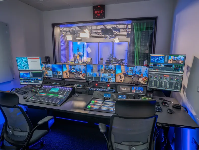 StoneX's production gallery, with an audio desk, vision mixer and multiviewer screens on a height-adjustable desk, and a window into the studio.