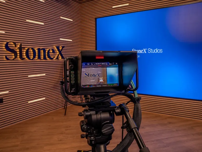 A studio camera on a tripod in StoneX's video studio, framing the backlit StoneX logo on a wood-slat wall next to a screen reading StoneX Studios.