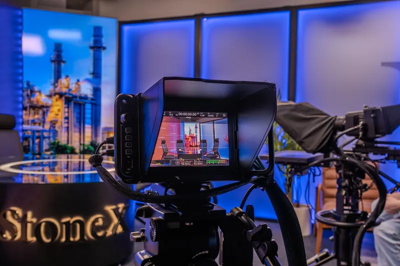 A studio camera viewfinder framing the round presenter table in StoneX's video studio, with the illuminated StoneX desk logo beside it.