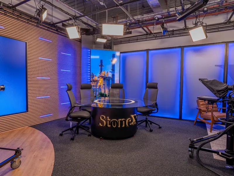 The round presenter table in StoneX's video studio, surrounded by chairs, blue backlit panels and an overhead LED lighting rig, with a pedestal camera and prompter on the right.