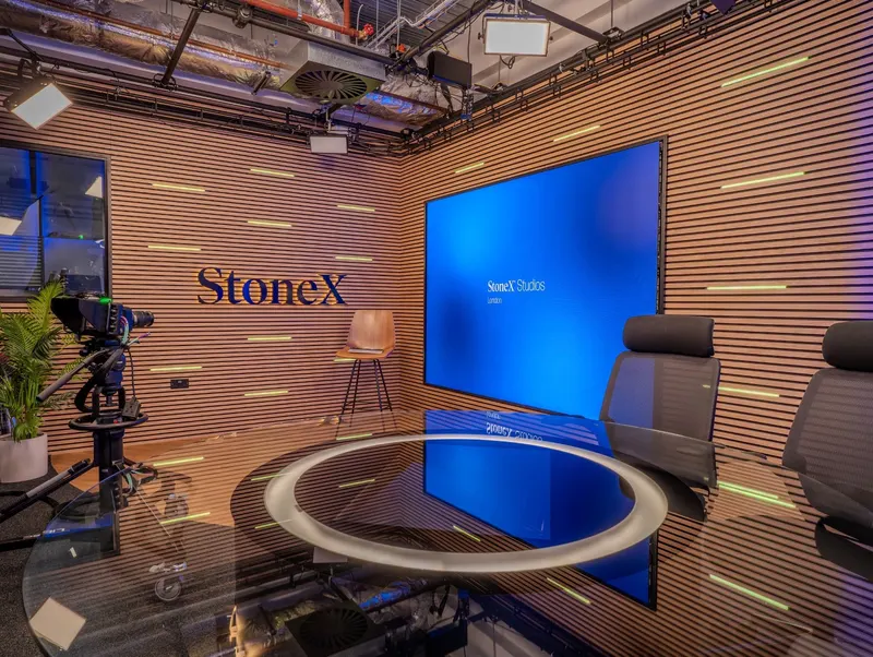 StoneX's video studio, with a round glass presenter table in front of a wood-slat wall carrying the StoneX logo and a large screen reading StoneX Studios London.